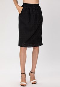 Jupe noire de longueur genou avec une taille élastique, présentant un tissu texturé et des découpes décoratives à l'ourlet, ainsi que des poches latérales.