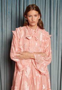 Mladá žena s dlhými vlasmi stojí pred modrými závesmi, oblečená v ružových kvetovaných šatách s volánikmi, mašľovým golierom a perlovými náušnicami.