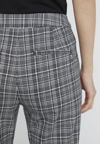 Detailní záběr na osobu v černobílých kostkovaných kalhotách s zadní kapsou, stojící na jednobarevném šedém pozadí.