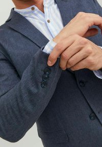 Pánsky blejzer z tmavomodrej látky. Má klopový golier, detaily na tri gombíky na manžetách a štruktúrovaný povrch. Nosený cez pruhovanú košeľu.