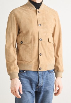 Heren beige suède bomberjack met knoopsluiting en klepzakken, gedragen over een zwart shirt en blauwe jeans.