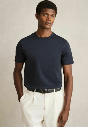 Tricou scurt cu mânecă, de culoare bleumarin, cu guler rotund, realizat dintr-un amestec de bumbac. Asortat cu pantaloni deschiși la culoare și o curea neagră texturată.