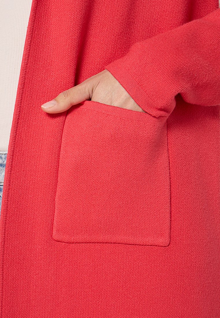 Roter Cardigan mit strukturiertem Strick, offener Front und einer aufgesetzten Tasche vorne. Die Ärmelenden sind leicht tailliert.