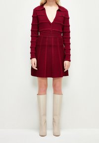 Robe tricotée rouge avec un décolleté en V profond, texture côtelée et fermeture à boutons, associée à des bottes beiges montantes. Présente des lignes structurées.