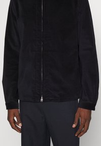 Giacca in corduroy nera con una texture liscia, chiusura frontale con zip e una vestibilità leggermente ampia. Maniche lunghe con polsini cuciti.