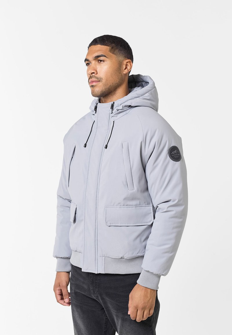 Grau gepolsterte Jacke mit Kapuze, ausgestattet mit zwei Vordertaschen und elastischen Bündchen. Logodetail am linken Ärmel. Glatte Textur.