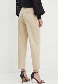 Pantaloni in cotone beige con design affusolato, dotati di tasche posteriori e una texture liscia. Abbinati a sandali neri con tacco alto.