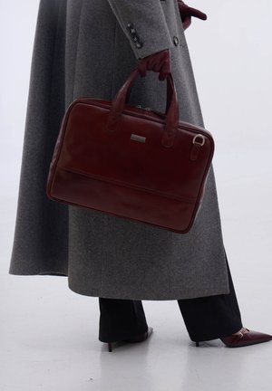 Personne en manteau gris et pantalon noir portant une mallette en cuir rouge foncé, avec des gants rouge foncé assortis et des chaussures à bout pointu.