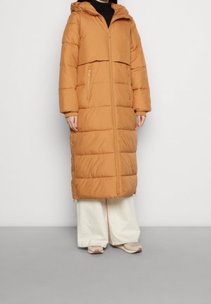 Person wearing a long, tan hooded puffer coat, white wide-leg pants, and beige sneakers standing against a plain light background.