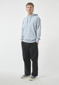 Light blue hoodie with a front pocket, drawstring hood, and small logo. Paired with black pants and blue sneakers, standing against a white background.