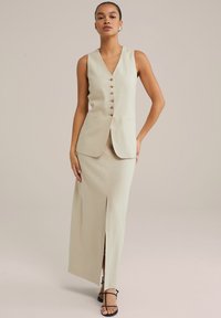 Beige mouwloos vest met een V-hals en vijf knopen, gecombineerd met een lange rok met een split aan de voorkant, gemaakt van textuurstof.