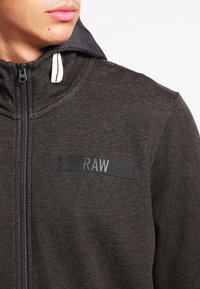 Felpa grigia con zip, caratterizzata da un'etichetta nera "RAW" sul petto, tessuto testurizzato e fodera interna del cappuccio di colore contrastante.