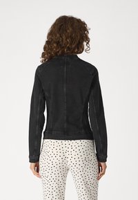 Osoba s kudrnatými vlasy, oblečená v upnuté černé džínsové bundě a bílých kalhotách s černými puntíky, stojící na jednobarevném pozadí.