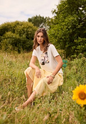 Jeune femme aux cheveux ondulés portant un t-shirt graphique et une jupe jaune, assise sur une chaise verte dans un champ herbeux près d'un tournesol.