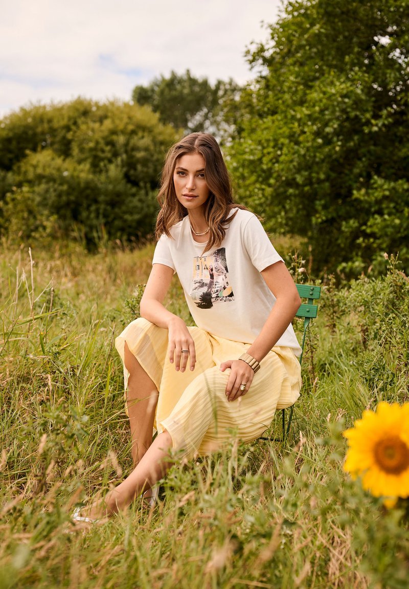 Jeune femme aux cheveux ondulés portant un t-shirt graphique et une jupe jaune, assise sur une chaise verte dans un champ herbeux près d'un tournesol.