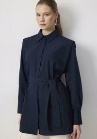 Námornícky modrá košeľa na gombíky s golierom, dlhými rukávmi a opaskom na viazanie v páse. Hladká textúra a priliehavý strih so bočnými rozparkami na spodnom okraji.