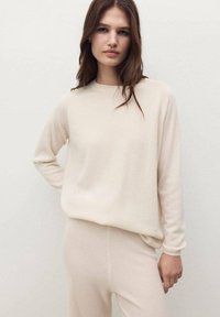 Pull en tricot beige avec un col rond et des manches longues, arborant une coupe décontractée et une texture douce, associé à des leggings assortis.