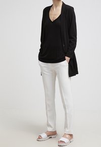 Persona vestida con un blazer negro y blusa, pantalones blancos y sandalias abiertas blancas, de pie con las manos en los bolsillos frente a un fondo liso.