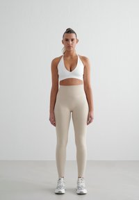 Witte halter sport-bh met uitsnede, gecombineerd met beige hoge taille legging en grijze sportschoenen, tegen een neutrale achtergrond.