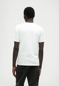 Biela bavlnená tričko s krátkymi rukávmi, okrúhlym výstrihom a hladkou textúrou, nosené s tmavými šedými džínsy; pohľad zozadu.