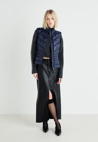 Navy padded vest with a zip front over a dark gray knit sweater, paired with a black leather skirt featuring a front slit, and black heels.