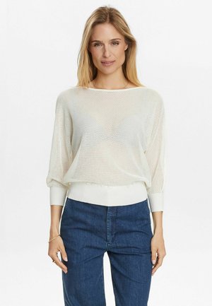 Weißer Netzpullover mit lockerer Passform, langen Ärmeln und gerippten Bündchen. Getragen über dunkelblauen Hosen mit einem strukturierten Strickmuster.
