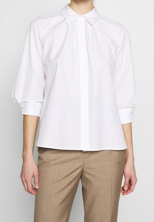Vrouw draagt een witte blouse met lange mouwen en kraag en beige broek, getoond van de nek tot halverwege de dijen tegen een effen achtergrond.