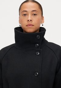 Melnas vilnas mēteles ar augstu apkakli un trim melnām pogām, kas izceļas ar pielāgotu piegriezumu un teksturētu audumu. Redzamas nav kabatas.