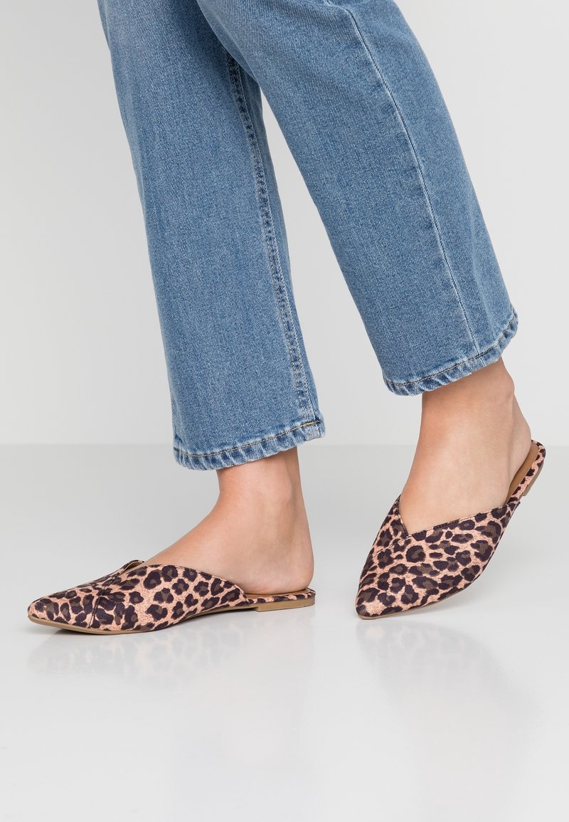 Pieds portant des ballerines pointues à imprimé léopard associées à un jean denim bleu coupé contre un fond uni.