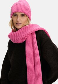 Różowy zestaw czapki i szalika z dzianiny, o prążkowanej fakturze. Szalik owinięty wokół szyi, a czapka dopasowana. Pod spodem czarny sweter.