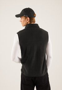 Black fleece vest with a high collar, worn over a white long-sleeve shirt. The model also wears a black cap, viewed from the back.