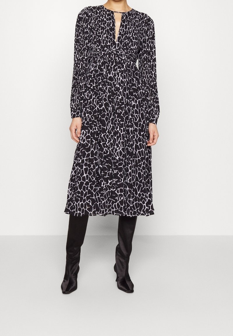 Femme portant une robe midi à manches longues à motifs noir et blanc associée à des bottes noires mi-mollet sur un fond uni.