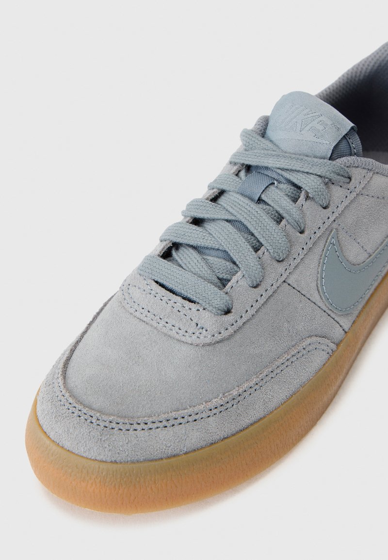 Zapatilla de ante gris con punta redondeada, detalles de cuero suave, cordones en gris claro y suela de goma. Detalle del logo cerca de la lengüeta.