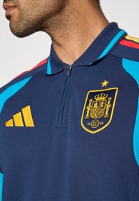Námornícka atletická polokošeľa s polovičným zipsom, s modrými a červenými prvkami, žltými pásmi Adidas a zlatým vyšivaným erbom Španielska.