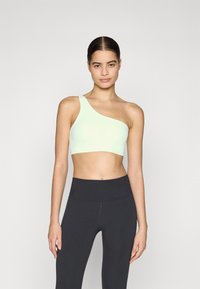 Sujetador deportivo asimétrico verde claro con una sola tirante, textura suave, combinado con leggins negros de cintura alta, diseño minimalista.