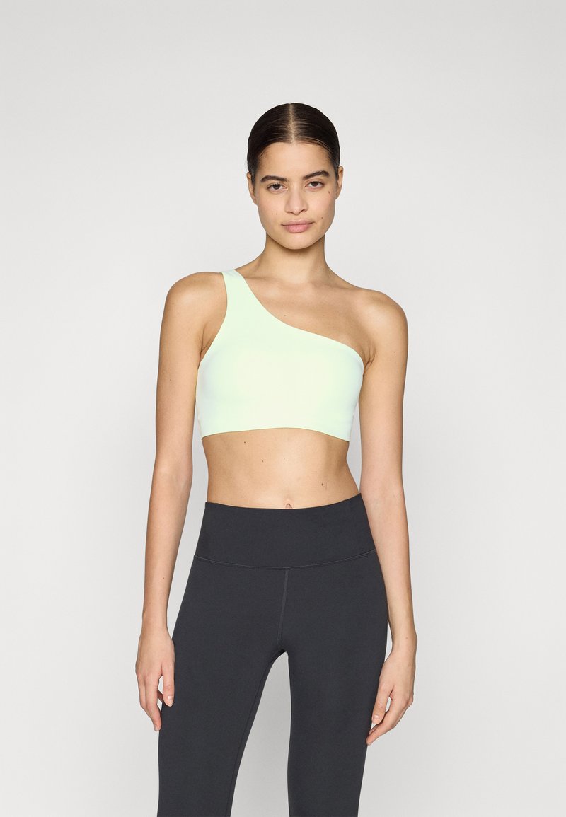 Sujetador deportivo asimétrico verde claro con una sola tirante, textura suave, combinado con leggins negros de cintura alta, diseño minimalista.