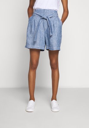 Persoon draagt lichtblauwe shorts met hoge taille en riem, wit T-shirt en witte sneakers, staand tegen een effen lichte achtergrond.