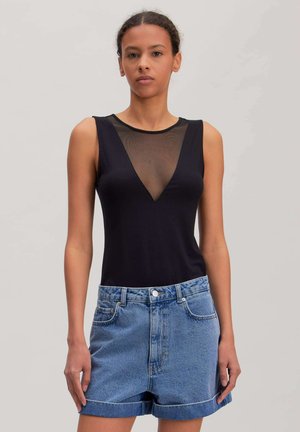 Vrouw draagt een mouwloos zwart topje met doorschijnende V-hals en lichtblauwe, hooggesneden, opgerolde denimshorts, staand tegen een effen achtergrond.