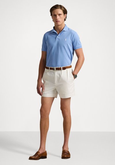 Cămașă polo bleu deschis, pantaloni scurți albi, curea maro și mocasini maronii. Cămașa polo are un guler clasic și un logo pe piept.
