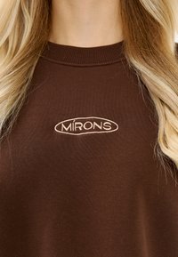 Brązowa bluza z okrągłym dekoltem z beżowym logo "MIRONS" umieszczonym na środku klatki piersiowej, noszona przez osobę z długimi blond włosami.