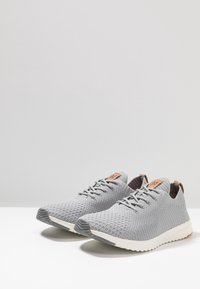 Scarpe atletiche a maglia grigia chiara con superficie testurizzata, dettagli in pelle beige sulla linguetta e suola leggera bianca.