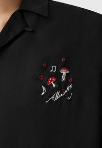Musta paita, jossa on brodeerattuja sieniä, musiikkimerkkejä ja punaisia sydämiä. "Allsaints"-logo on ommeltu valkoisena. Silkkinen kangas.