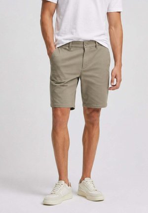 Uomo con pantaloncini beige fino al ginocchio, maglietta bianca e scarpe da ginnastica bianche, con le mani nelle tasche, in piedi su sfondo semplice.