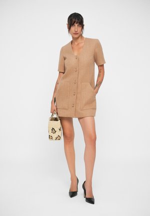 Μακρύ μανίκι καφέ φόρεμα με κουμπιά και δύο μπροστινές τσέπες, συνδυασμένο με μια πλεκτή τσάντα χειρός με μαύρο μοτίβο και μαύρες ψηλές γόβες.