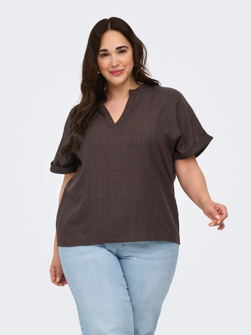 Femme aux longs cheveux foncés portant un haut ample à manches courtes marron foncé et un jean bleu clair, souriante sur un fond clair.