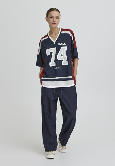 Femme portant un maillot de sport bleu marine et bordeaux avec le numéro 74, un pantalon bleu marine ample et des baskets blanches, devant un fond clair uni.