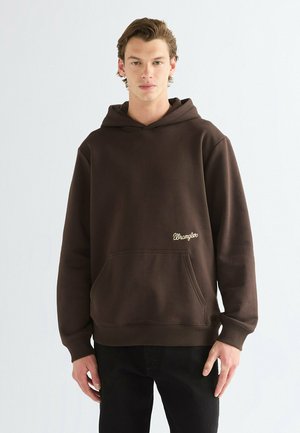 Junger Mann trägt einen dunkelbraunen Wrangler Hoodie mit Fronttasche und schwarzen Hosen, steht vor einem schlichten hellen Hintergrund.