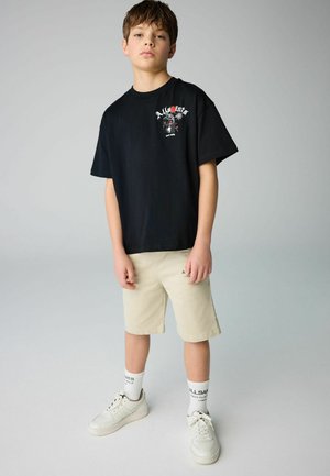 T-shirt nera in cotone con stampa grafica sul petto, abbinata a pantaloni corti color khaki. Sneakers bianche e calzini alti completano l'outfit.