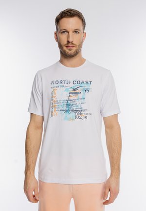 Mężczyzna o krótkich włosach i brodzie, ubrany w białą koszulkę z nadrukiem o tematyce morskiej oraz jasnoróżowe spodnie, stojący na białym tle.