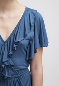 Turkusowa sukienka z głębokim dekoltem w kształcie litery V i warstwowymi rękawami z falbanami. Miękka tkanina o gładkiej fakturze, wcięcie w talii zapewniające dopasowany krój.